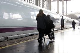 Una persona en silla de ruedas se dirige a un tren, acompañada por una ayudante